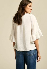 Witte getextureerde blouse met geribbelde mouwen, gecombineerd met blauwe denim jeans. De stof heeft een subtiele kreukeleffect, wat visuele interesse creëert.