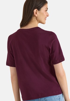 Žena s hnědými vlasy, která má na sobě tmavě fialový krátký rukáv s průstřihy na lemech rukávů, pohled zezadu.