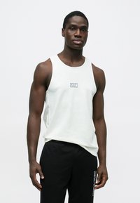 Mladý muž v bielom tielku EA7 a čiernych nohaviciach Emporio Armani stojí na jednoduchom svetlom pozadí.