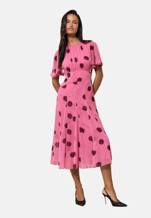 Frau trägt ein rosa Midi-Kleid mit schwarzen Punkten, kurzen Flatterärmeln und schwarzen High Heels, steht vor einem einfarbigen Hintergrund.