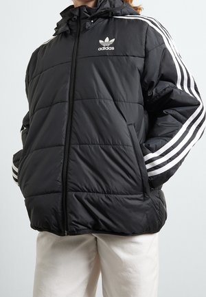 Personne portant une doudoune noire Adidas avec des bandes blanches sur les manches et le logo sur la poitrine, associée à un pantalon de couleur claire.