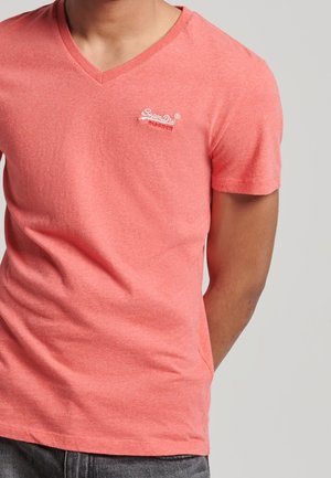 Homme portant un T-shirt rose saumon à col en V et manches courtes avec un logo "SuperDry" blanc brodé sur la poitrine, pantalon gris, sur fond uni.