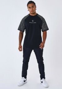 Camiseta negra con mangas grises estampadas y texto "PROJECT X PARIS". Combinada con pantalones azul marino y zapatillas blancas con detalles negros.