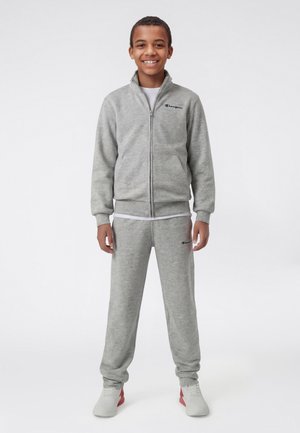 Ragazzo sorridente in piedi che indossa una giacca grigia con zip e pantaloni della tuta abbinati, con scarpe da ginnastica bianche, su sfondo bianco.
