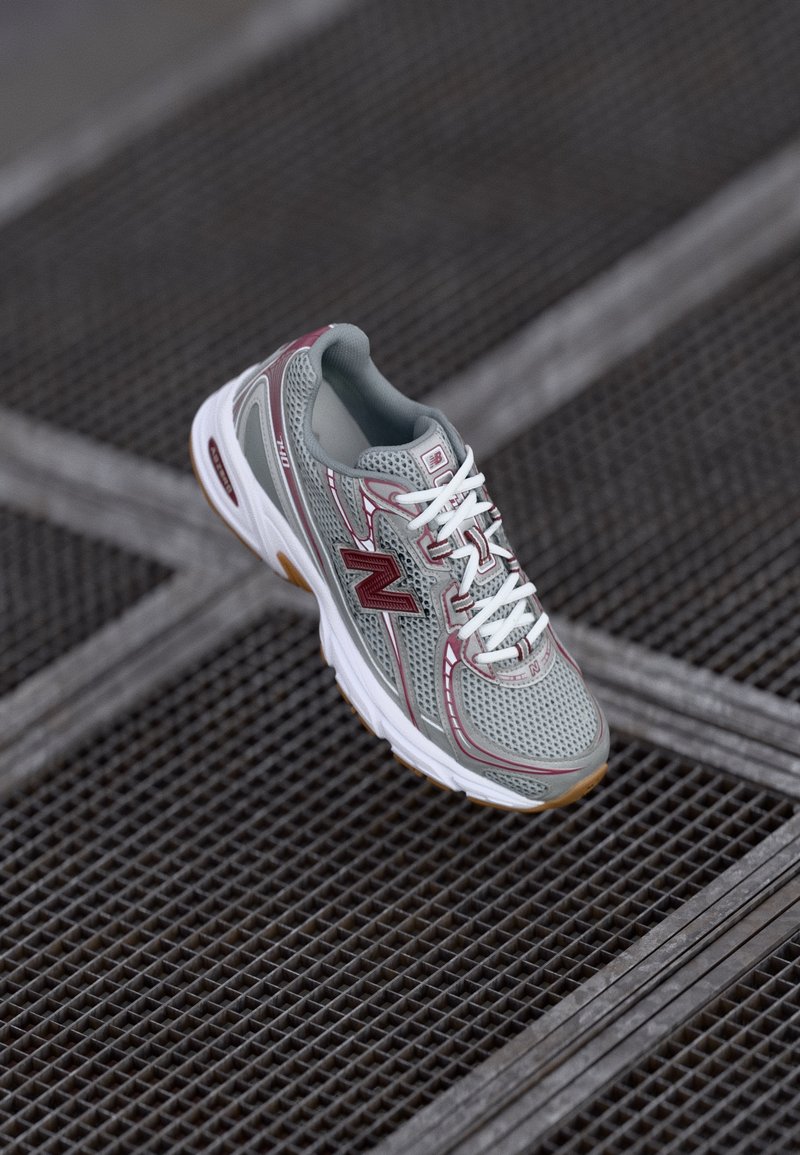 Chaussure de course grise et bordeaux avec des lacets blancs reposant sur une surface en grille métallique sombre avec des motifs carrés.