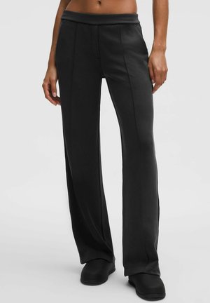 Sort bukser med bred benvidde i en glat stof, elastisk talje og diskrete forsejlede sømme, båret med sorte slip-on sko.