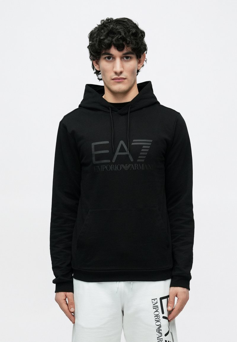 Mladý muž s kučeravými vlasmi, oblečený v čiernej mikine EA7 Emporio Armani a bielych teplákoch s čiernym logom Emporio Armani, stojaci pred bielym pozadím.