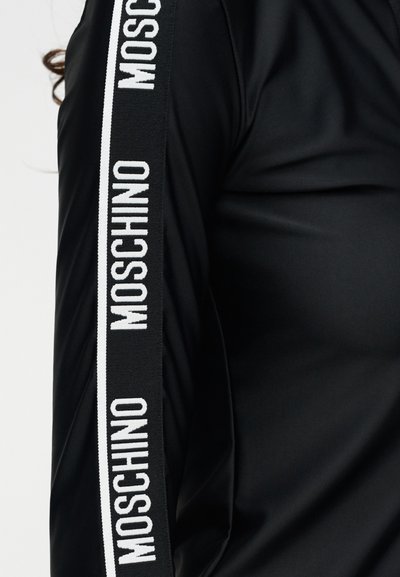 Μαύρο τοπ με μακριά μανίκια με λευκό κείμενο «MOSCHINO» επαναλαμβανόμενο κατά μήκος της εξωτερικής ρίγας του μανικιού.