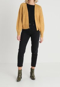 Cardigan lavorato a maglia giallo senape con apertura frontale e maniche lunghe, abbinato a jeans neri a vita alta e stivaletti verde verniciato.