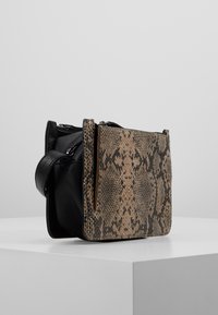 Una borsa elegante caratterizzata da un corpo in pelle nera con una patta a motivo rettile, esposta su un piedistallo bianco.