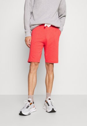 Persona che indossa una felpa grigia, pantaloncini color corallo rossi fino al ginocchio, calzini marrone chiaro e scarpe da ginnastica bianche, in piedi davanti a uno sfondo chiaro uniforme.