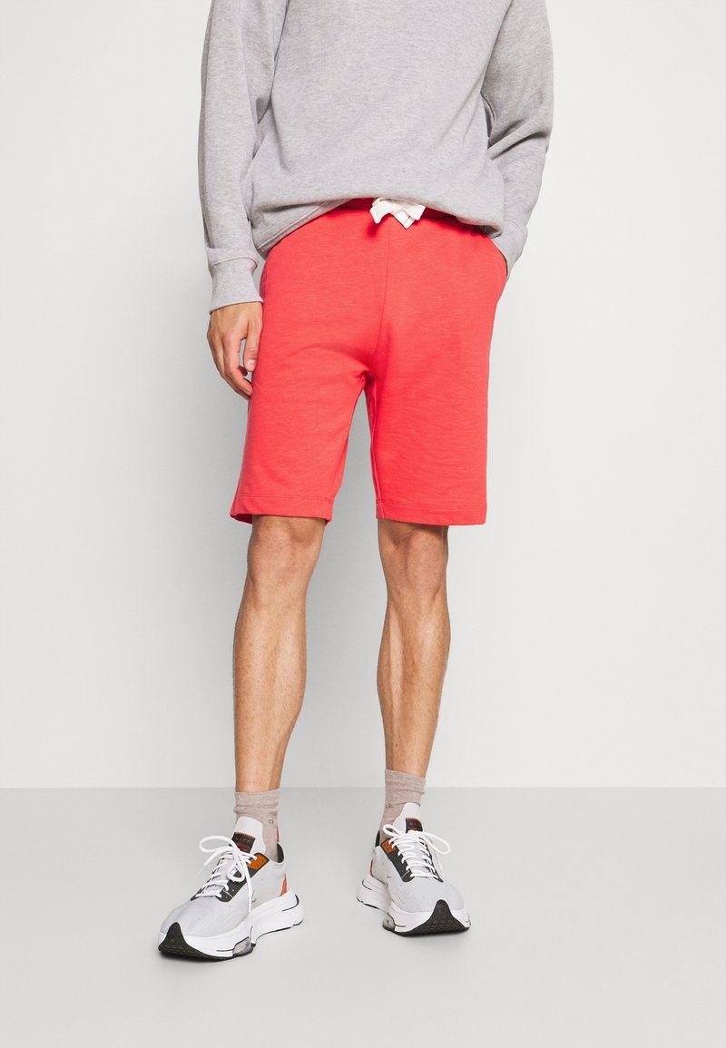 Personne portant un sweat-shirt gris, un short corail rouge aux genoux, des chaussettes marron clair et des baskets blanches, debout devant un fond clair uni.