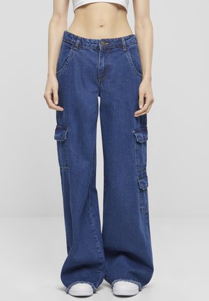 Frau trägt weite blaue Denim-Cargohosen mit Klappentaschen und weiße Sneakers, steht vor einem schlichten hellen Hintergrund.