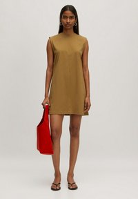 Kurzärmliges, olivgrünes Kleid mit hohem Kragen, flacher Textur und minimalen Nähten. Kombiniert mit einer leuchtend roten Tragetasche und schwarzen Sandalen.