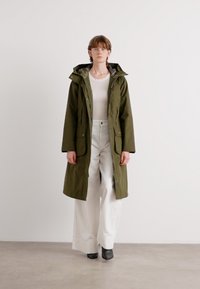Olijfgroene, middenlange parka met een capuchon, voorzien van twee voorzakken en een rits, gedragen over een witte top en wijde broek.