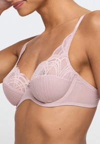 Biustonosz z bieliźniarskim w miękkim różu, z koronkowymi detalami, formowanymi miseczkami oraz regulowanymi ramiączkami. Zawiera dekoracyjny metalowy akcent z przodu.
