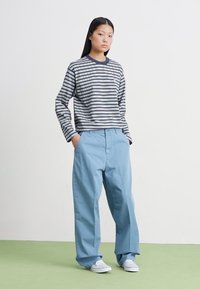 Striped long-sleeve shirt in navy and white, paired with light blue wide-leg pants. Model wears white sneakers, standing on a green surface.