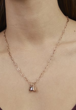 Collier chaîne en or rose avec maillons allongés, orné de deux pendentifs : un petit bijou violet et un charme de teinte claire, reposant sur la clavicule.