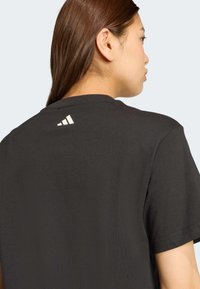 Juodas marškinėliai su apykakle ir trumpomis rankovėmis. Prie nugaros apykaklės yra baltas Adidas logotipas. Lygus audinio paviršius.