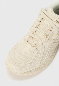 Baskets de sport beige avec panneaux en maille et en suède, lacets épais et semelle texturée, vues d'un angle avant supérieur.