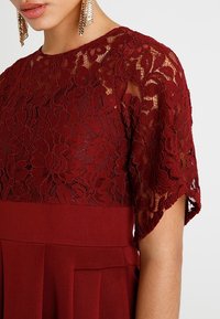 Burgundy klänning med spetsöverdrag som har blomm önster, korta ärmar och en figursydd bodice, kompletterad med en plisserad kjol.