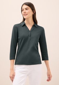 Tmavě zelená, dlouhý rukáv, polo tričko s límečkem, se zapínáním na tři knoflíky a hladkou texturou, kombinované s bílými kalhotami.