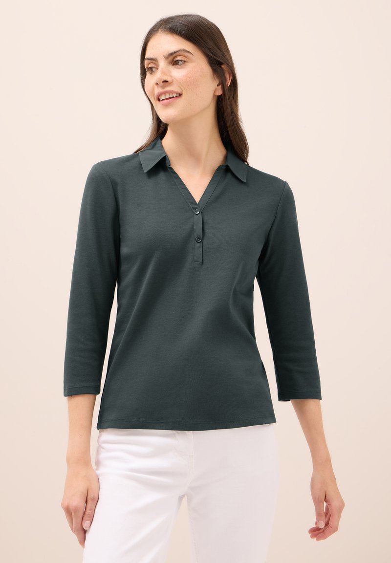 Tmavě zelená, dlouhý rukáv, polo tričko s límečkem, se zapínáním na tři knoflíky a hladkou texturou, kombinované s bílými kalhotami.