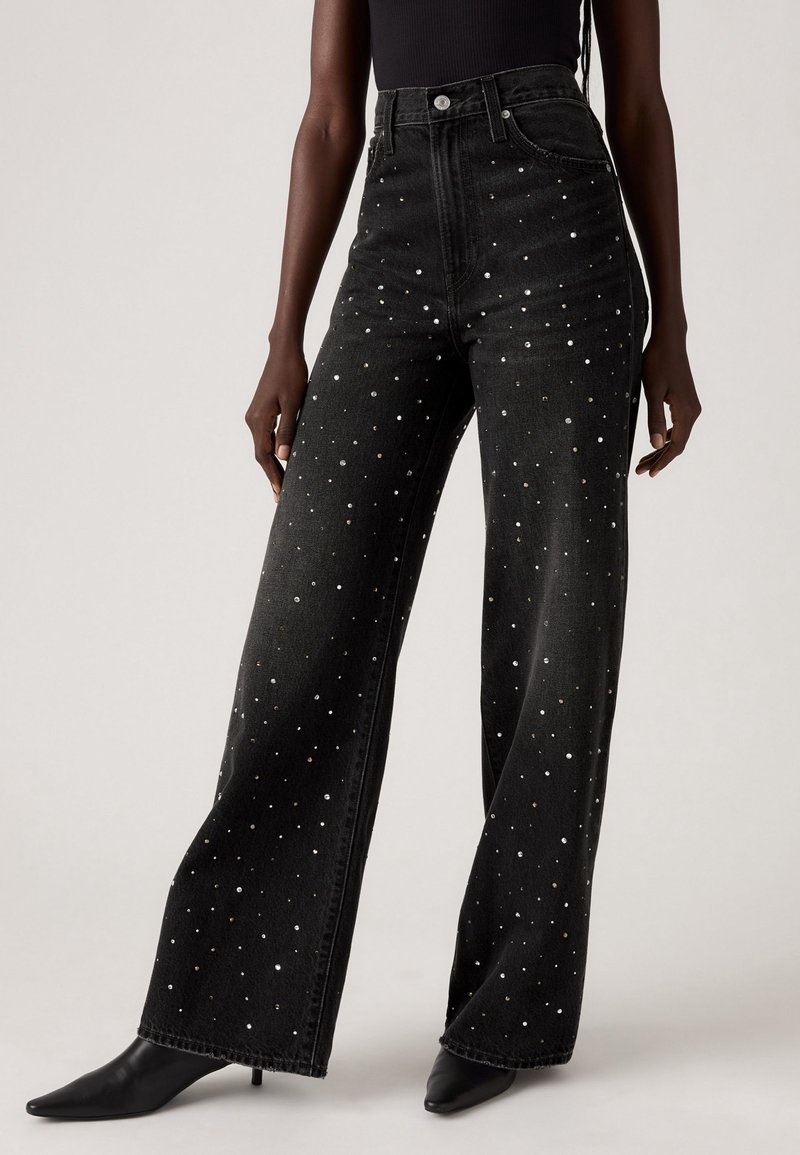 Jean noir taille haute à jambes larges avec des clous argentés dispersés, porté avec des chaussures noires à bout pointu par une personne debout.