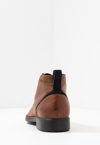 Brun läderankelboot med en mockakrage, mörkblå accent nära hälen och en strukturerad svart gummisula. Bakvy visas.