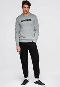 Mężczyzna ubrany w szarą bluzę z napisem "OMBRE", czarne spodnie cargo, białe skarpetki i czarne trampki, stojący na jednolitym tle.