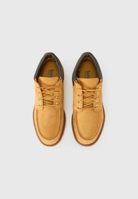 Lichtbruine suède schoenen met gele veters, rubberen zolen en een gevoerde donkergroene kraag. Deze schoenen hebben gestikte details langs de randen.