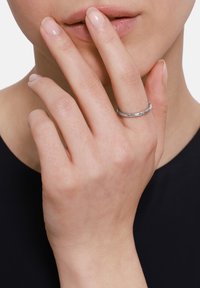 Srebrna pierścionek o teksturowanym, skręconym wzorze. Noszony na smukłej ręce, palce delikatnie dotykają ust, na ciemnym tle.