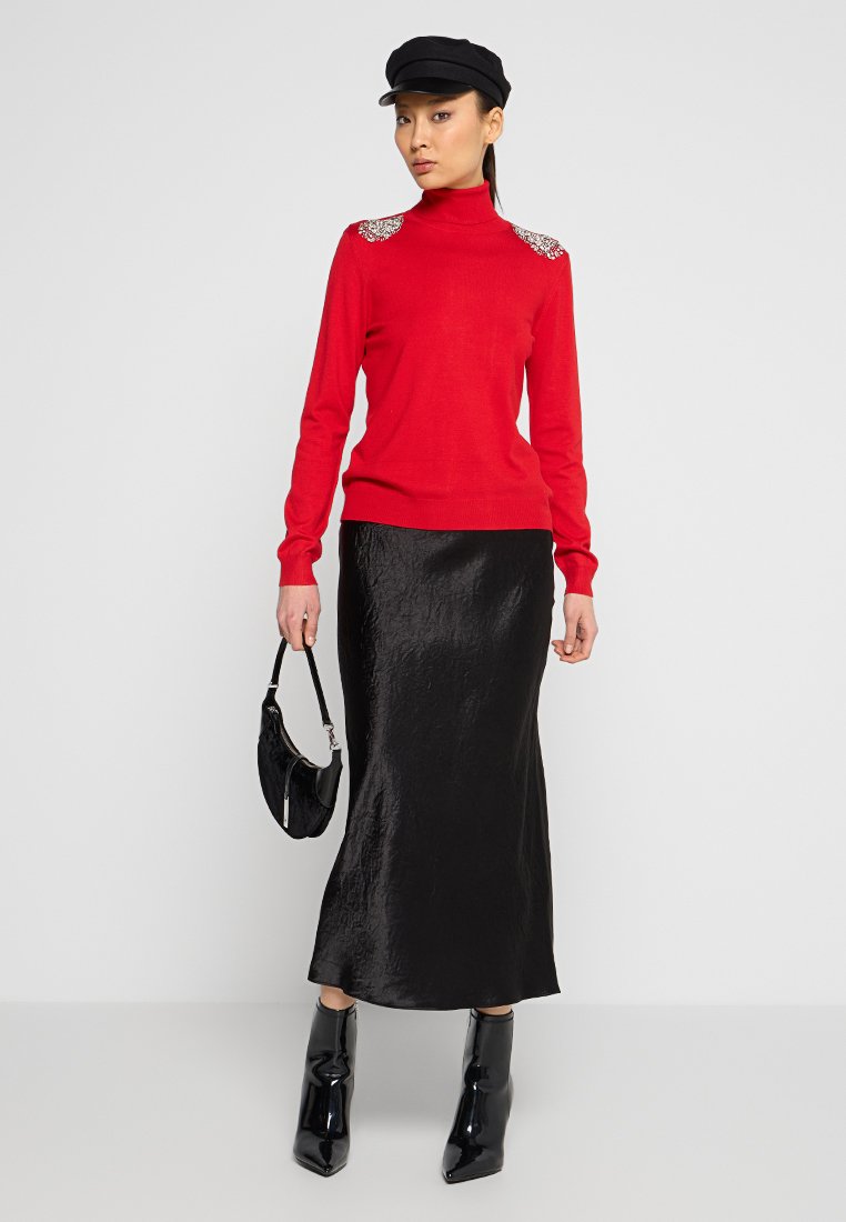 Maglione rosso a collo alto con decorazioni metalliche sulle spalle, abbinato a una gonna midi nera in satin e stivali alla caviglia neri lucidi. Borsa nera.