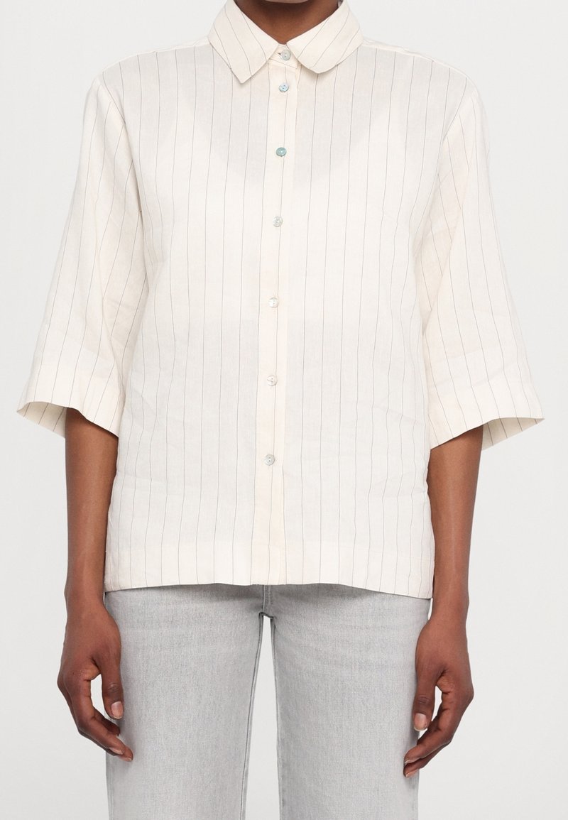 Chemise à col crème avec des rayures verticales bleu marine, manches courtes et fermeture boutonnée à l'avant avec des boutons bleu clair. Tissu léger.