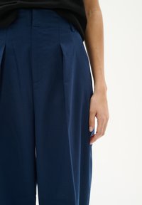 Marinefarbene, weit geschnittene Hosen aus glattem Stoff, mit Falten an der Vorderseite, einem taillierten Bund und einer seitlichen Tasche entlang der Naht.