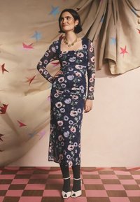 Ilgomis rankovėmis, aptempta suknelė su gėlių raštu tamsiai mėlynos ir rožinės spalvų tonais. Suknelėje yra kvadratinis iškirptės dizainas ir raukiniai, derinami su juodomis ribinėmis kojinėmis ir baltais aukštakulniais.