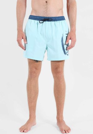 Homme portant un short de bain bleu clair avec une ceinture bleu marine et un cordon noir, debout pieds nus sur un fond blanc.