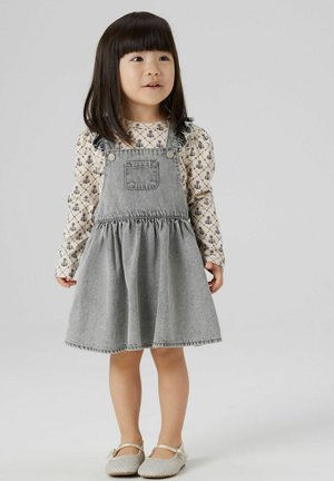 Grå denimkjole med flossede stropper, samlet talje og udvidet nederdel; lagdelt over en hvid langærmet bluse med blomsterprint.