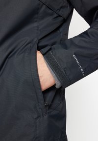 Black waterproof jacket with a textured finish, featuring a side pocket, velcro cuff adjustment, and "OMNI-TECH" branding on the sleeve.