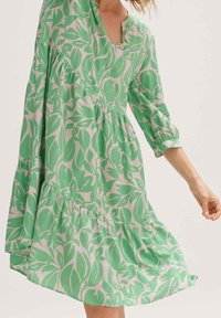 Kobieta ubrana w luźną zieloną i beżową sukienkę w liście z rękawami do łokci i marszczonymi szwami na jasnym tle.
