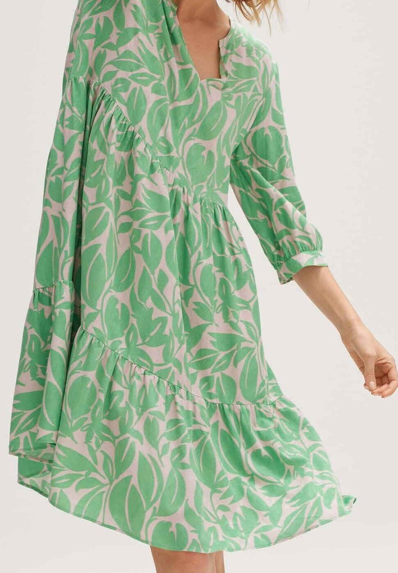 Kobieta ubrana w luźną zieloną i beżową sukienkę w liście z rękawami do łokci i marszczonymi szwami na jasnym tle.