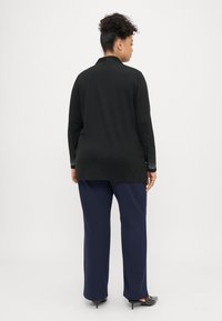 Μαύρο μακρυμάνικο πουκάμισο με διακοσμητικά στις μανσέτες, συνδυασμένο με σκούρες μπλε παντελόνι. Η υφή είναι λεία, με κλασικό γιακά και χαλαρή εφαρμογή.