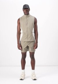 Débardeur sans manches de couleur beige clair avec des accents blancs, associé à un short assorti. Porté avec une casquette noire et des chaussures de sport beiges.