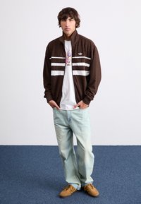 Καφέ ζακέτα με φερμουάρ και υφασμάτινη υφή με λευκές ρίγες, συνδυασμένη με ανοιχτό �μπλε τζιν και καφέ αθλητικά παπούτσια. Λευκό μπλουζάκι από κάτω με γραφικά.