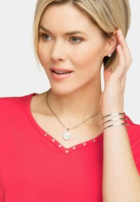 Rode v-hals top met zilveren studs, voorzien van een zilveren ketting met een octagonaal hangertje en gestapelde zilveren armbanden om de pols.