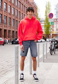 Ljusrosa hoodie med känguruficka, kombinerat med ljusblå denimshorts med fransade kanter och ett hål vid knät. Svart- och vitrandiga sneakers.