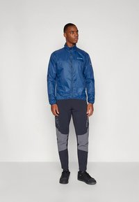 Tumši zils viegls jaka ar rāvējslēdzēja aizdari, kopā ar tumšām biksēm ar pelēkiem akcentiem. Modelis stāv melnās apavos.