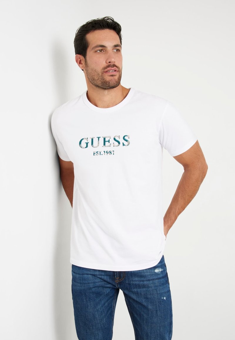 T-shirt blanc à col rond arborant le logo "GUESS" en bleu et un design texturé, associé à un jean en denim bleu. Tissu lisse, coupe décontractée.
