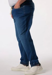 Pantalones de mezclilla azul oscuro con cintura elástica, cierre de cordón, dobladillos con puños y bolsillos laterales, combinados con zapatillas blancas.
