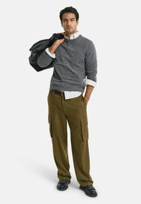 Pull en tricot gris par-dessus une chemise à col blanc, pantalon cargo olive avec poches latérales et chaussures noires. Un sac noir est porté sur une épaule.